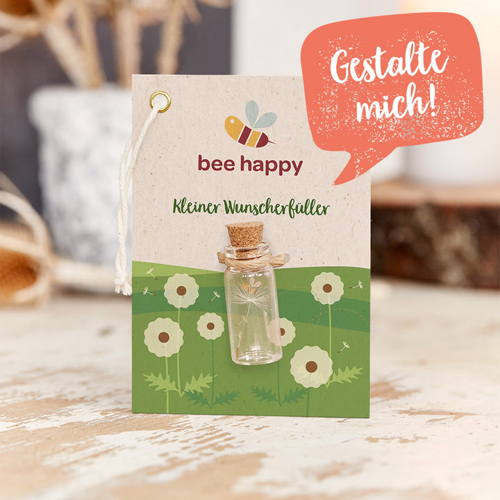 Carte avec l'inscription "bee happy" et "Little wish-exaucer", une petite bouteille en verre attachée, un arrière-plan avec une prairie dessinée et des pissenlits, et une bulle de dialogue avec le texte "Design me!".