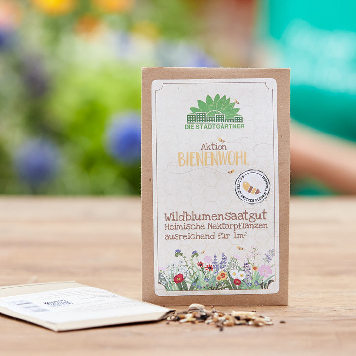 Sac en papier avec des graines de fleurs sauvages « Aktion Bienenwohl » sur une table en bois, fleurs colorées floues en arrière-plan.