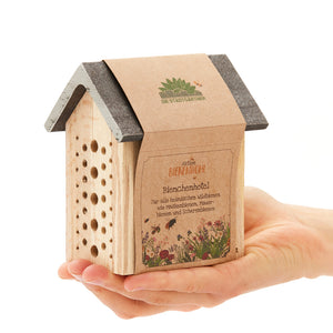 Une main tient un petit hôtel à abeilles en bois avec l'inscription « Aktion Bienenwohl Bienenhotel » en allemand.