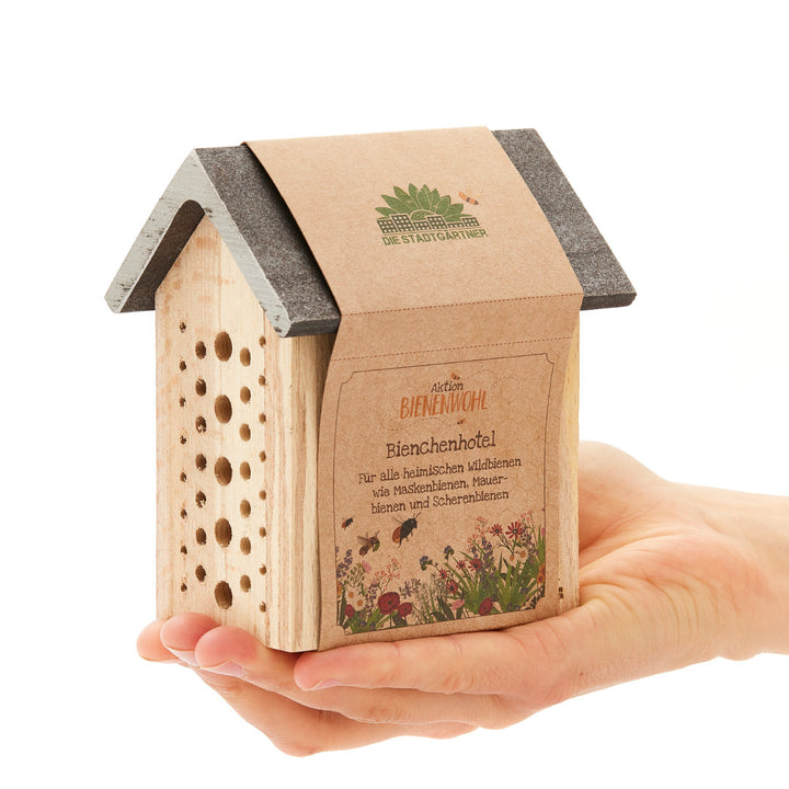 Une main tient un petit hôtel à abeilles en bois avec l'inscription « Aktion Bienenwohl Bienenhotel » en allemand.