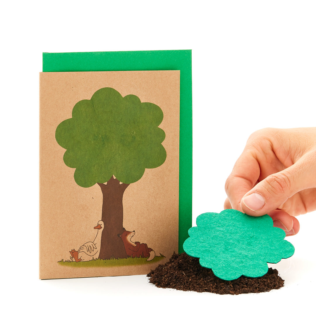 Une main tient une cime d'arbre en papier vert devant une carte de vœux avec un tronc d'arbre dessiné et des animaux sur fond blanc.