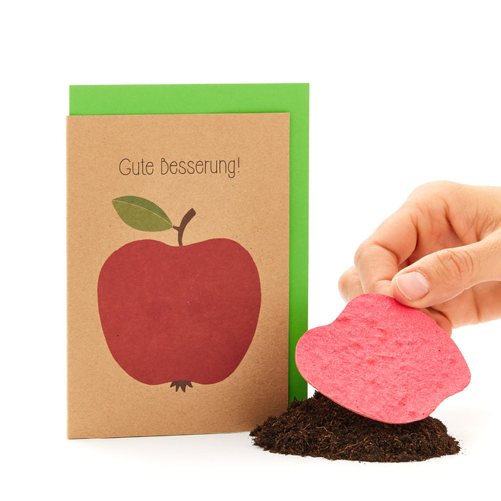 Une carte « bon rétablissement » avec une pomme dessus à côté d'une main plantant une carte de graines en forme de cœur dans le sol.