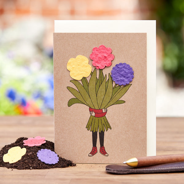 Carte de vœux faite à la main avec des jambes dessinées dans des chaussures rouges tenant un bouquet de fleurs en papier colorées sur une table en bois avec un stylo à côté.