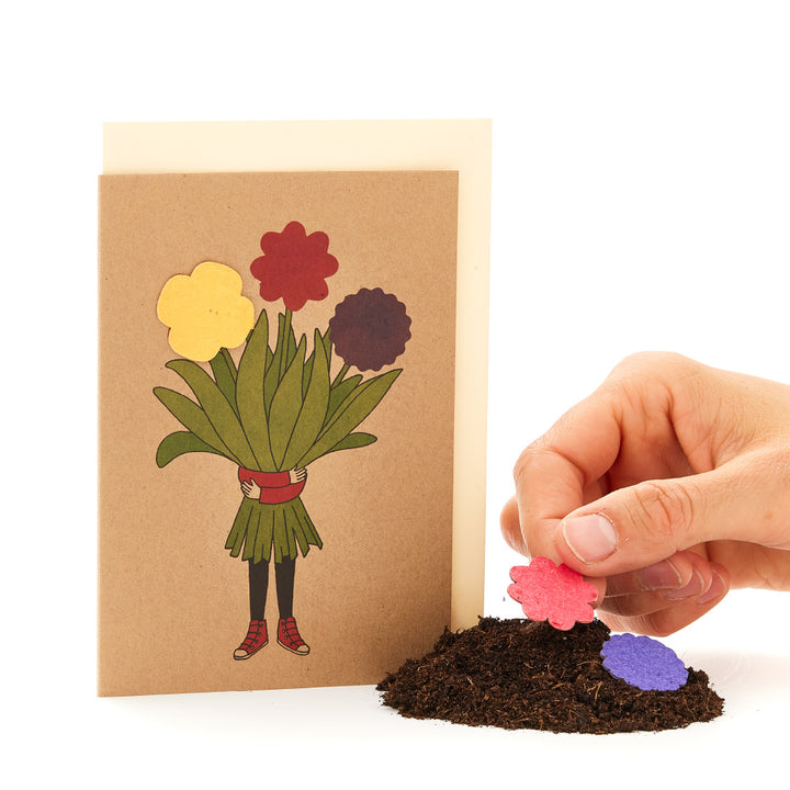 Main plaçant une fleur en papier sur un tas de terre à côté d'une carte de vœux avec un motif de bouquet.