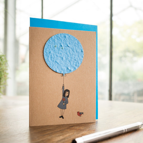 Carte de vœux faite à la main avec une figure dessinée flottant sur un ballon et un oiseau sur une table en bois.