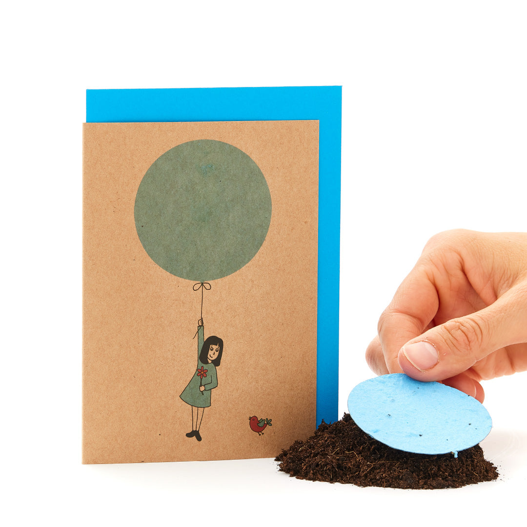Une carte de vœux avec une figure dessinée suspendue à un ballon et une main plaçant un papier ensemencé rond bleu sur le sol.