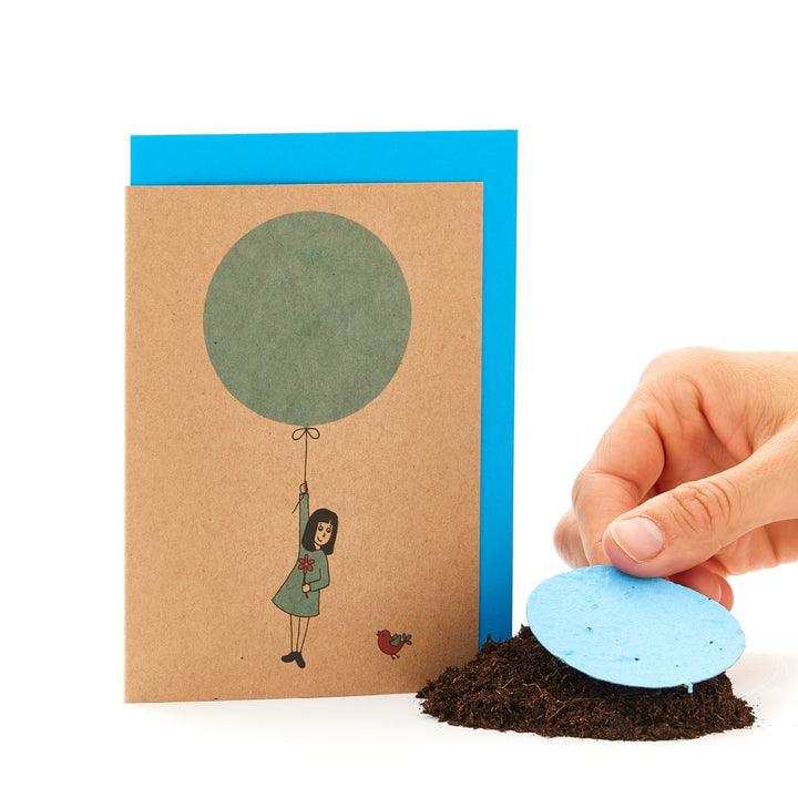 Une carte de vœux avec une figure dessinée suspendue à un ballon et une main plaçant un papier ensemencé rond bleu sur le sol.