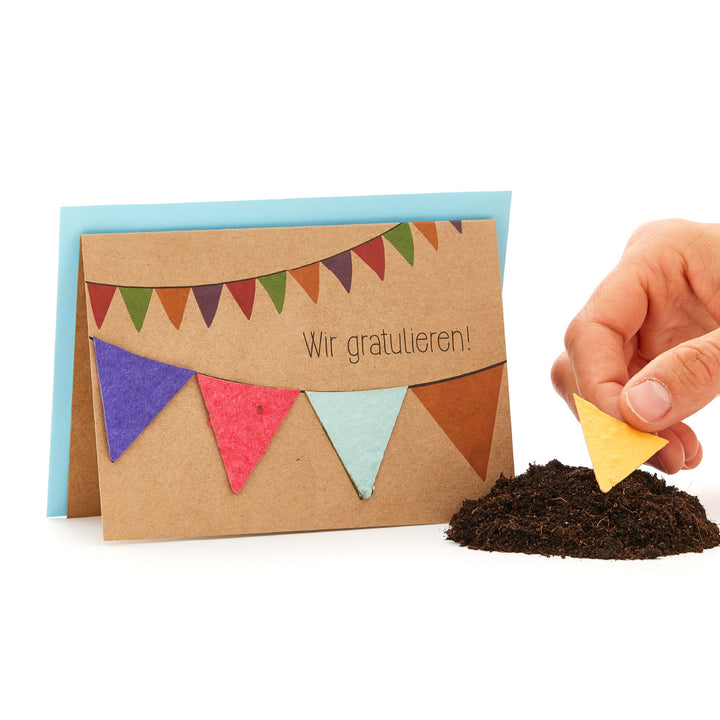 Carte de vœux avec banderoles colorées et l'inscription « Félicitations ! », main tenant un triangle jaune au-dessus d'un monticule de terre.