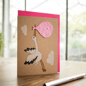 Carte de vœux avec une cigogne portant un paquet rose sur une table en bois avec un stylo à côté.