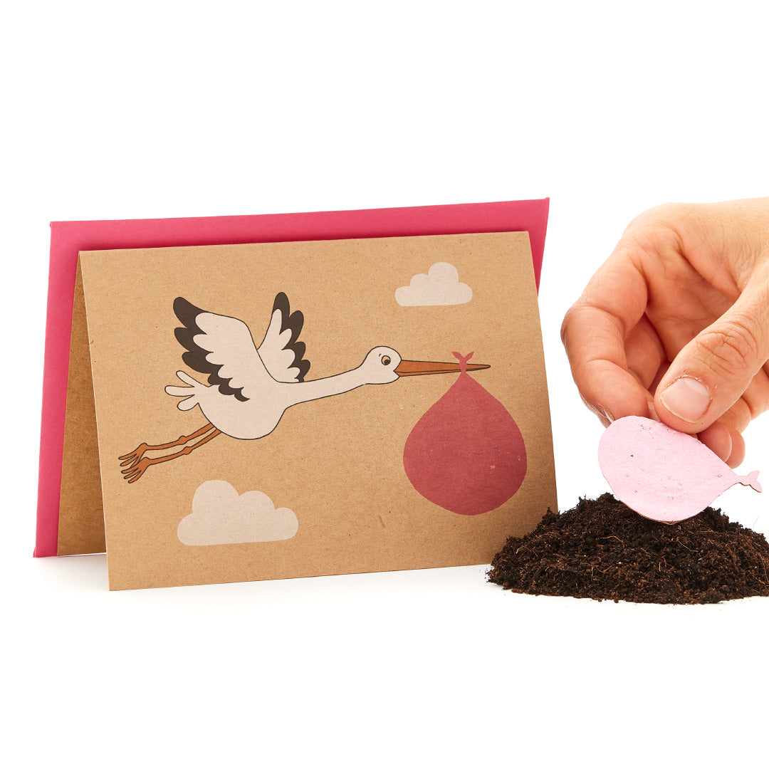 Cigogne sur une carte de vœux portant un paquet de bébés à côté d'une main tenant un objet rose en forme de cœur sur un tas de terre.