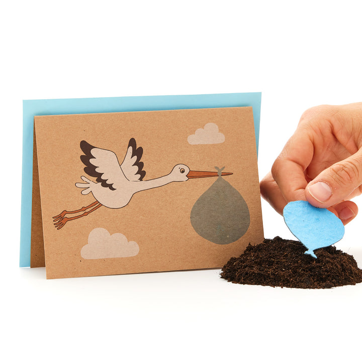 Carte de vœux avec une cigogne portant un paquet de bébés à côté d'une main plantant une graine bleue en forme de cœur dans le sol.
