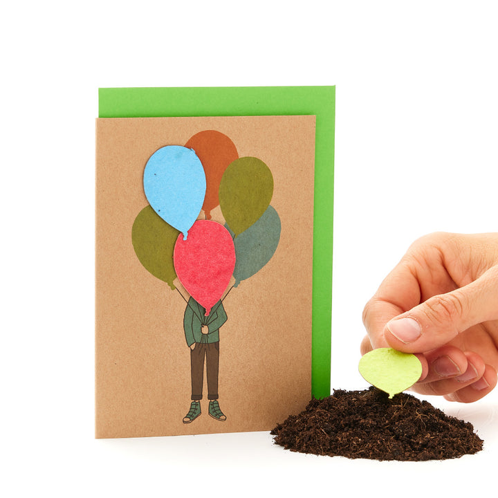 Carte de vœux avec un personnage illustré tenant des ballons à côté d'un tas de terre à partir duquel pousse un vrai ballon.
