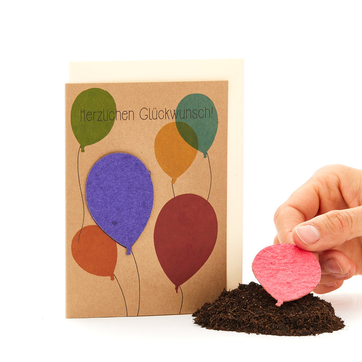 Carte de vœux avec des ballons colorés et les mots « Félicitations » à côté d'un tas de terre et d'une main plantant un ballon rose.