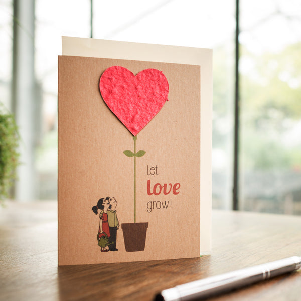 Carte de vœux avec un couple s'embrassant à côté d'un pot avec une plante qui en sort avec un cœur rouge, avec le texte "laissez l'amour grandir !"