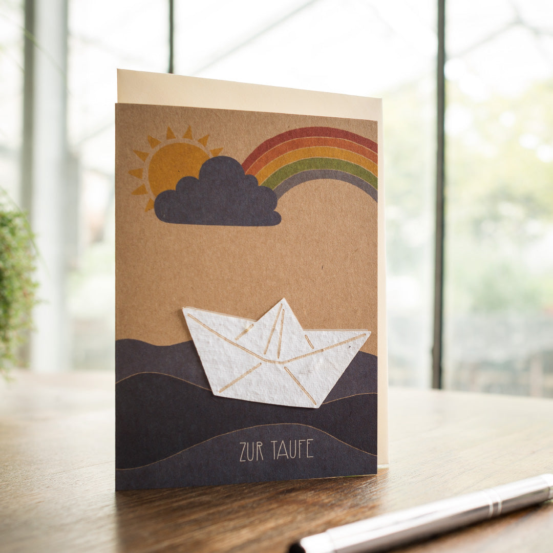 Carte de baptême avec bateau en papier et arc-en-ciel sur table en bois.