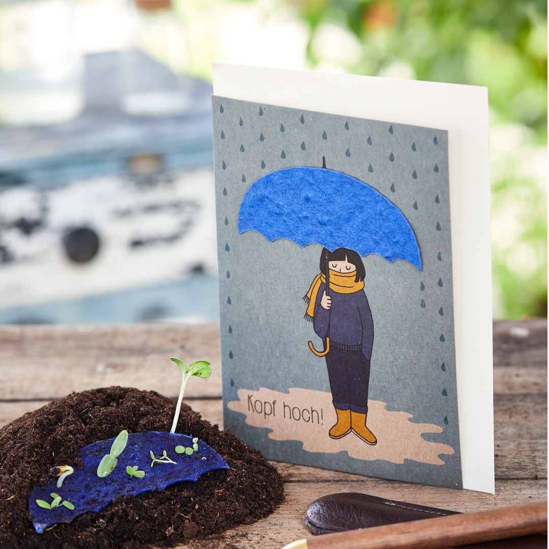 Carte de voeux avec un dessin d'une personne sous un parapluie bleu et l'inscription "Chin up!", à côté d'une jeune plante sur terre.