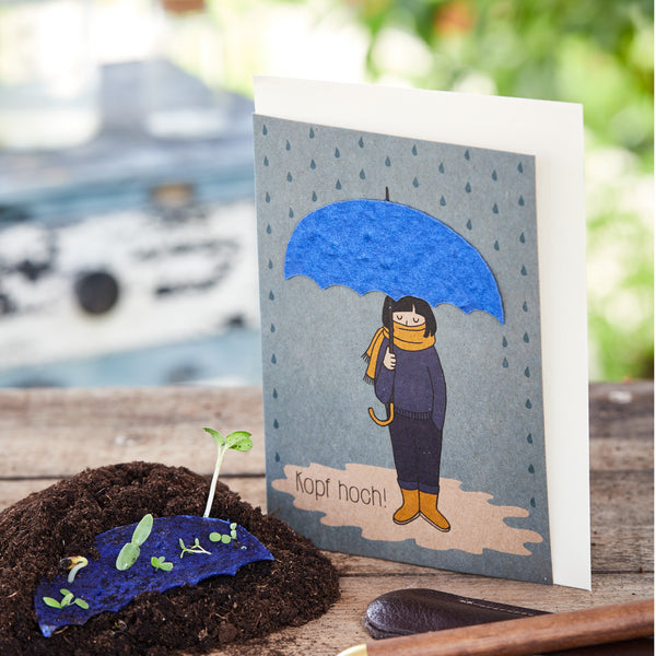 Carte de voeux avec un dessin d'une personne sous un parapluie bleu et l'inscription "Chin up!", à côté d'une jeune plante sur terre.