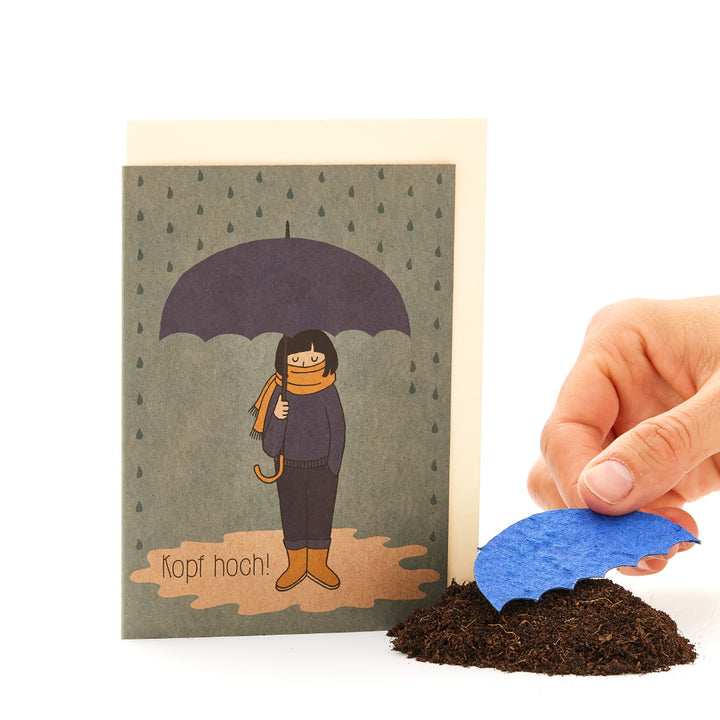 Main plaçant une vague de papier bleu sur un tas de terre devant une carte de vœux avec une silhouette dessinée sous un parapluie et le texte « Chin up ! »
