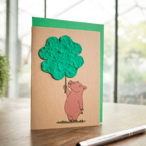 Carte de vœux faite à la main avec un cochon dessiné tenant un arbre en papier vert sur une table en bois avec un stylo.