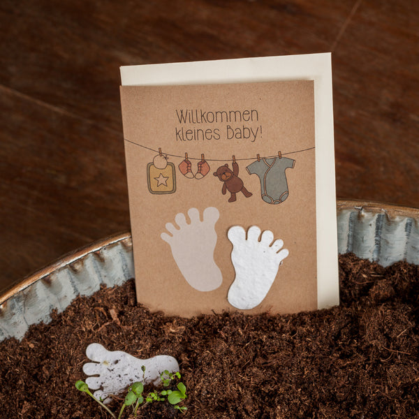 Carte de baby shower avec l'inscription "Bienvenue petit bébé !" et des illustrations de pieds de bébé, d'un cintre avec une barboteuse et de jouets, sur un fond de terre et de bois.