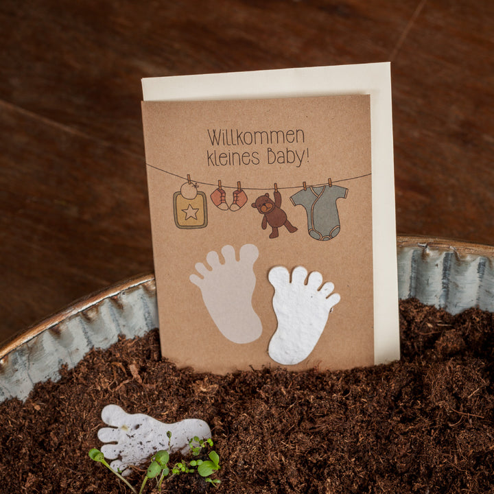 Carte de baby shower avec l'inscription "Bienvenue petit bébé !" et des illustrations de pieds de bébé, d'un cintre avec une barboteuse et de jouets, sur un fond de terre et de bois.