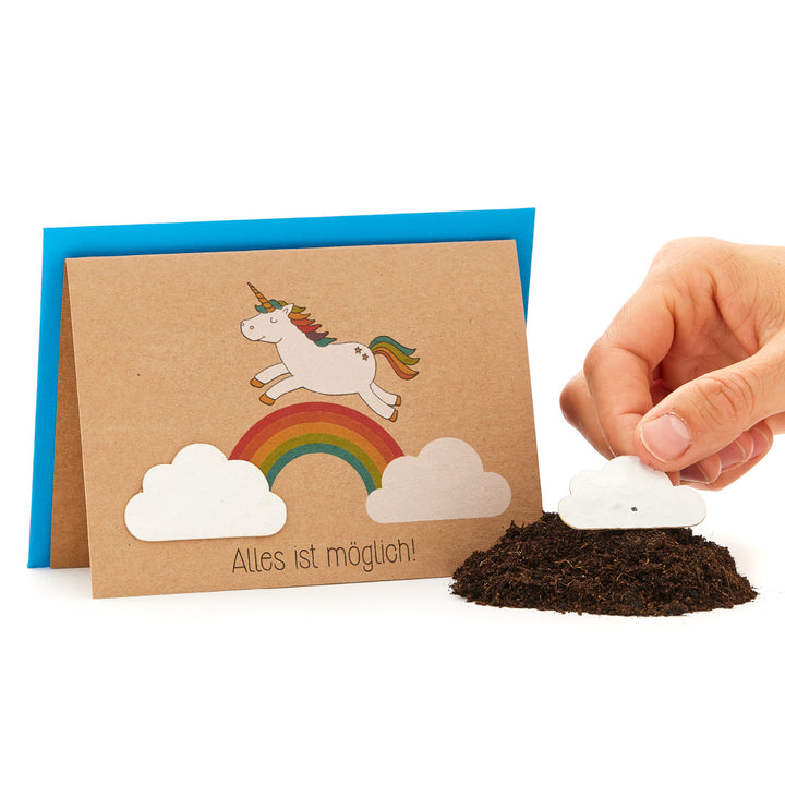 Une main plante une graine de papier en forme de nuage dans le sol, à côté d'une carte de vœux avec une licorne sur un arc-en-ciel et le dicton « Tout est possible ! ».
