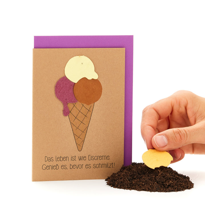 Main plantant un biscuit en forme de cœur dans la terre, à côté d'une carte de vœux en forme de cornet de glace avec le slogan : « La vie est comme une glace. Savourez-la avant qu'elle ne fonde ! »
