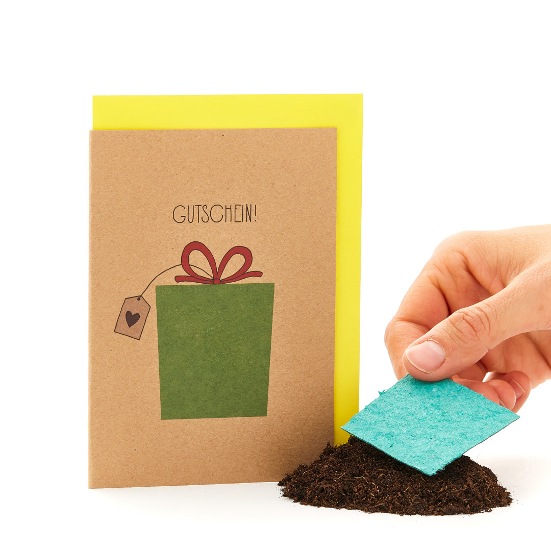 Main tenant une carte de vœux avec l'inscription "BON" et un cadeau dessiné, à côté d'un tas de terre avec un morceau de carton bleu.