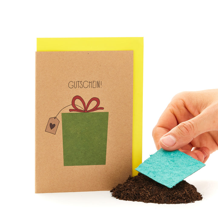 Main tenant une carte de vœux avec l'inscription "BON" et un cadeau dessiné, à côté d'un tas de terre avec un morceau de carton bleu.