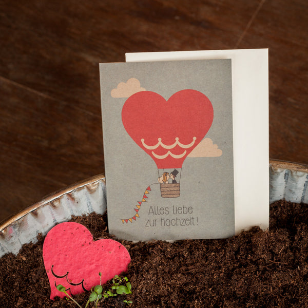 Carte de félicitations de mariage comportant une montgolfière en forme de cœur et l'inscription « Joyeux mariage ! », placée à côté d'un objet en forme de cœur et de terreau.