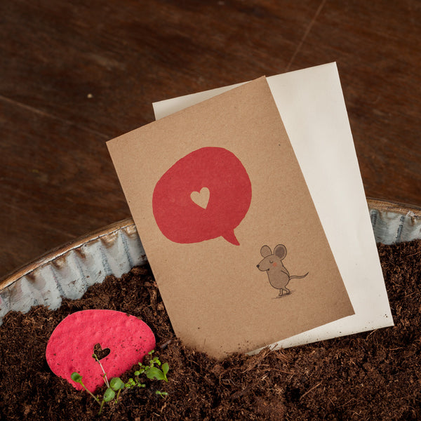 Une carte de vœux avec une souris dessinée et une bulle de dialogue rouge avec un cœur, à côté une enveloppe blanche et un cœur en papier rouge dans la terre.
