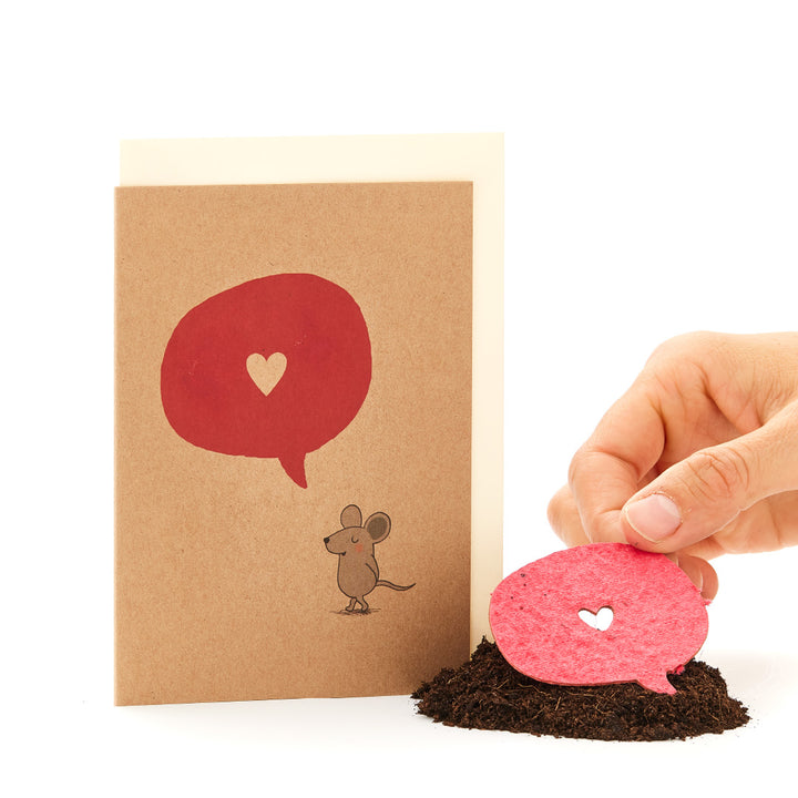 Une main plante une bulle de dialogue en papier rouge en forme de cœur dans le sol, à côté se trouve une carte avec une souris et une bulle de dialogue rouge.