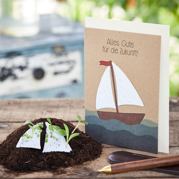 Carte de voeux avec un voilier et l'inscription "Tout le meilleur pour l'avenir!", à côté de la terre avec des semis et un stylo sur une table en bois.