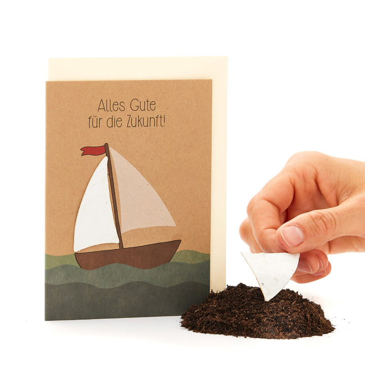 Carte de vœux avec un motif de voilier et l'inscription « Tous mes vœux pour l'avenir ! », à côté d'une main plantant un morceau de papier en forme de voile dans le sol.
