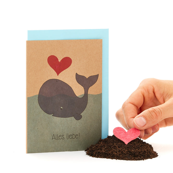 Carte de vœux avec une baleine imprimée et un cœur rouge, à côté d'elle une main plantant un cœur rouge dans le sol.