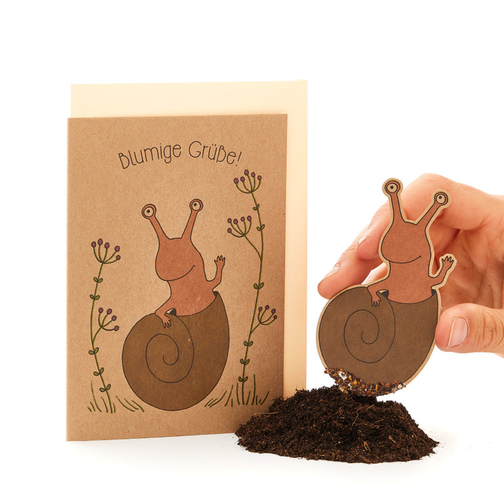Personne tenant une illustration en papier découpé d'un escargot sur de la vraie terre, assortie d'une carte de vœux avec le texte « Salutations fleuries ! » et un dessin d'escargot.