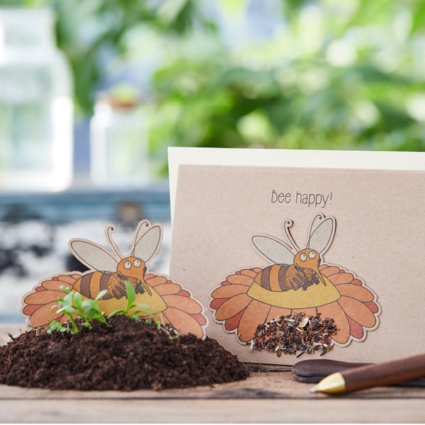 Bricolage avec un motif d'abeille et l'inscription "Bee happy!" sur une table avec de la terre et des plantes en arrière-plan.