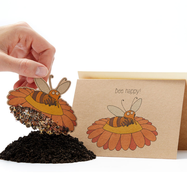 Une main tenant une carte de graines en forme d'abeille avec le slogan « Bee happy ! » dessus et un tas de terre en dessous.