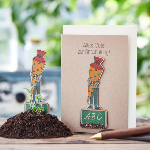 Carte de félicitations pour l'inscription à l'école avec une figurine de carotte et un tableau ABC sur une table en bois, entourée de plantes et de terre.
