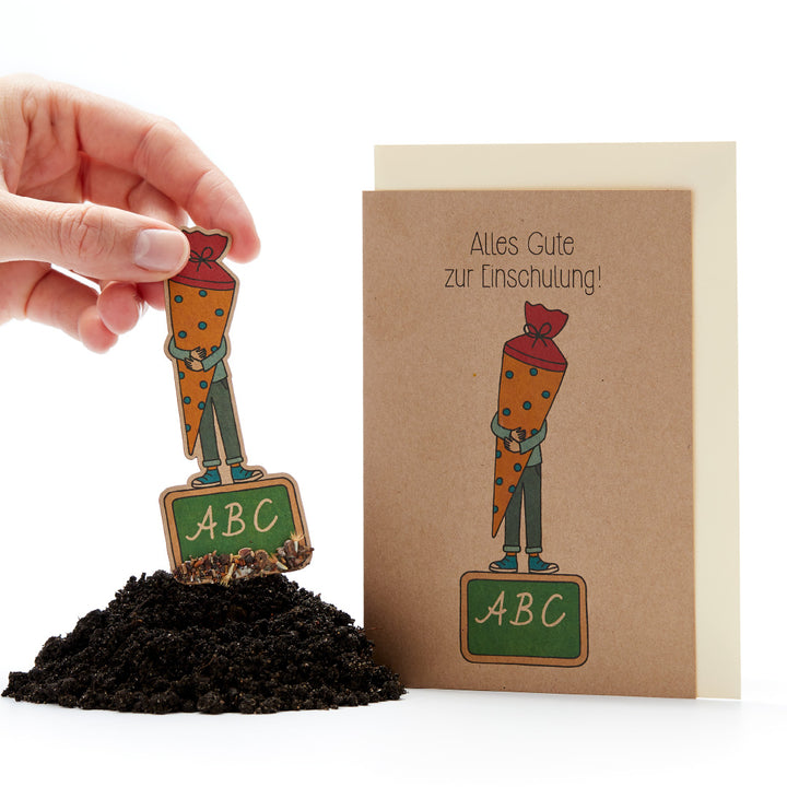 Main tenant une figurine de carotte avec une casquette d'école et un tableau ABC, à côté une carte de félicitations pour le premier jour d'école.