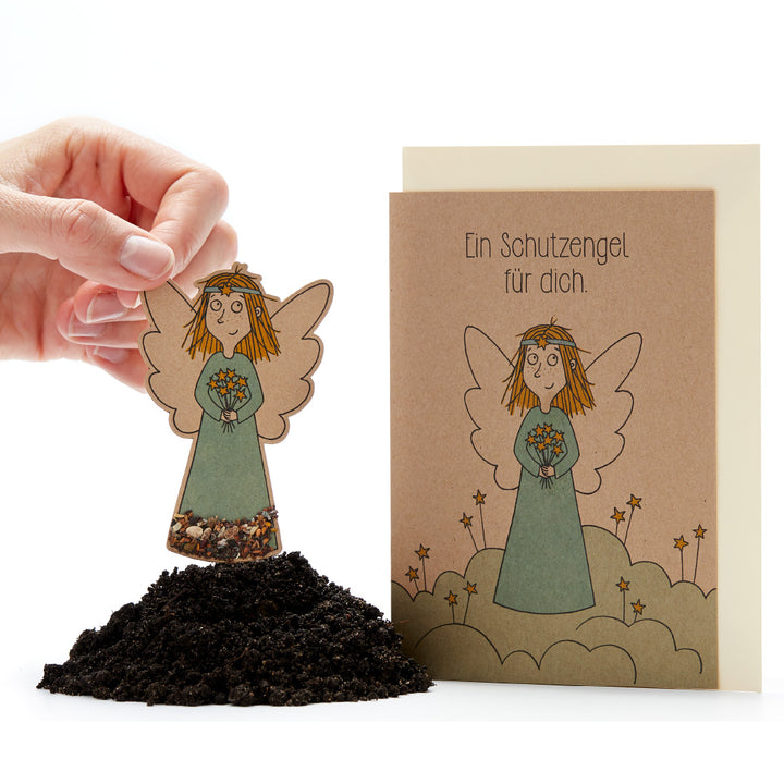 Main tenant une figurine en papier ensemencé d'un ange sur le sol à côté d'une carte avec le texte « Un ange gardien pour toi ».