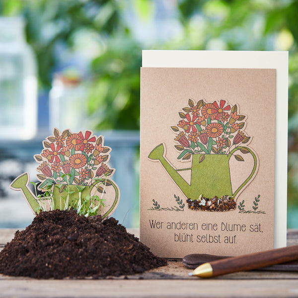 Carte de vœux avec une illustration d'un arrosoir rempli de fleurs et le dicton allemand « Celui qui sème une fleur pour les autres fleurira lui-même », à côté une carte de semis dans un pot de fleurs avec de la terre et de la verdure en herbe.