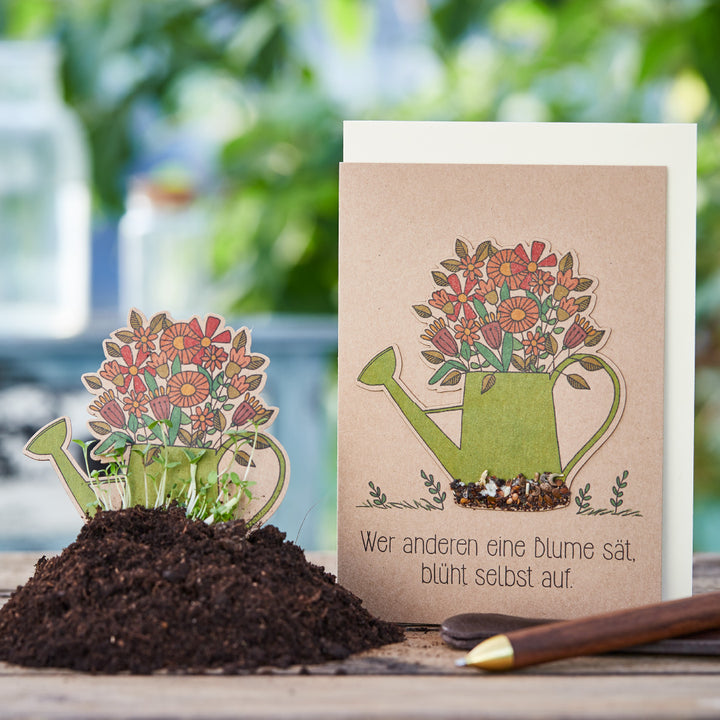 Carte de vœux avec une illustration d'un arrosoir rempli de fleurs et le dicton allemand « Celui qui sème une fleur pour les autres fleurira lui-même », à côté une carte de semis dans un pot de fleurs avec de la terre et de la verdure en herbe.