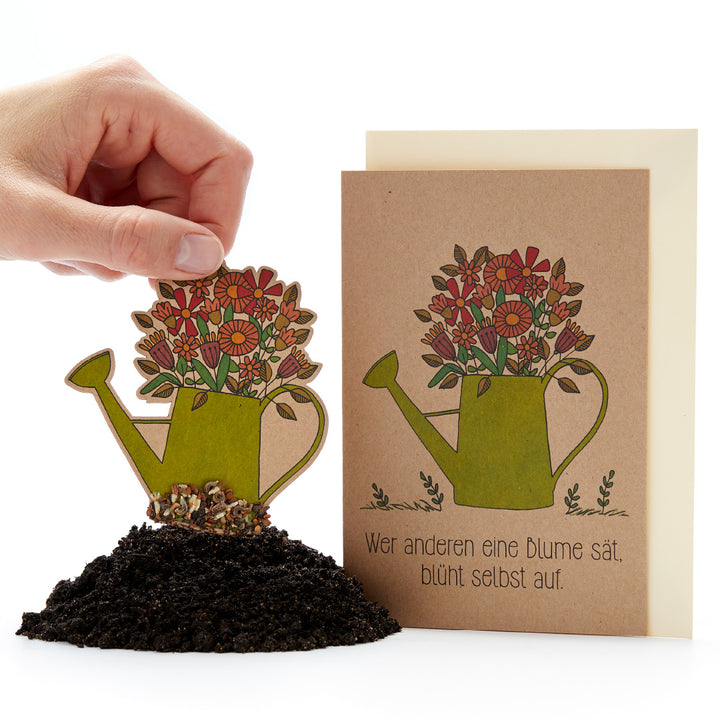 Une main place un arrosoir en papier décoré de fleurs dans la terre ; à côté se trouve une carte avec le même motif et le dicton allemand : « Celui qui sème une fleur pour les autres fleurira lui-même. »
