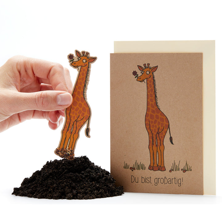 Une main plante une carte en forme de girafe dans le sol, à côté une carte de vœux avec un dessin de girafe et le texte "Tu es génial !".