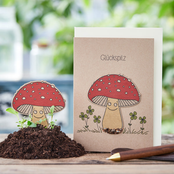 Carte de vœux avec des personnages de champignons de dessin animé et le mot « champignon porte-bonheur », à côté d'un vrai monticule de terre et d'un crayon.