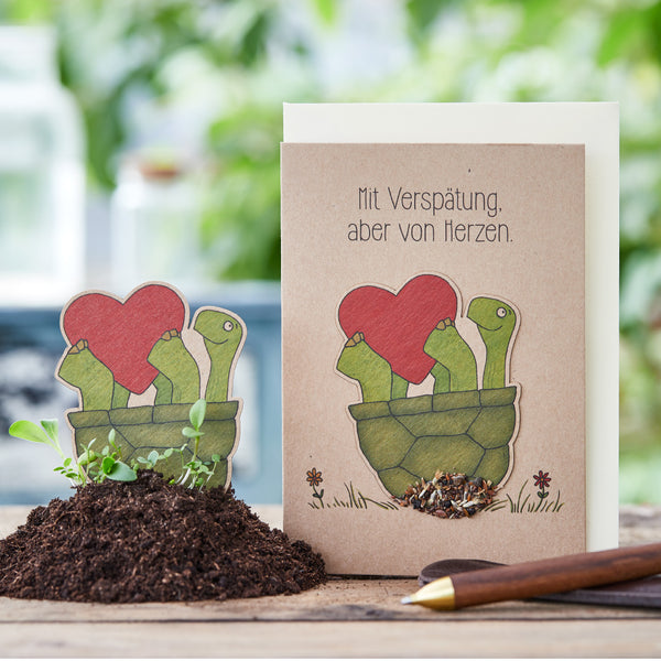 Carte de vœux représentant un dessin de tortues tenant un cœur rouge, avec un marqueur de plante assorti dans le sol à côté ; le texte sur la carte dit : « Retardé, mais du fond du cœur. »
