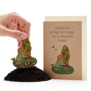 Une main plante une carte de graines représentant un jardinier et un plant de tomates dans le sol, à côté d'elle une carte de vœux sur laquelle on peut lire : « Le jardinage est moins cher que la thérapie, et vous obtenez des tomates ! »