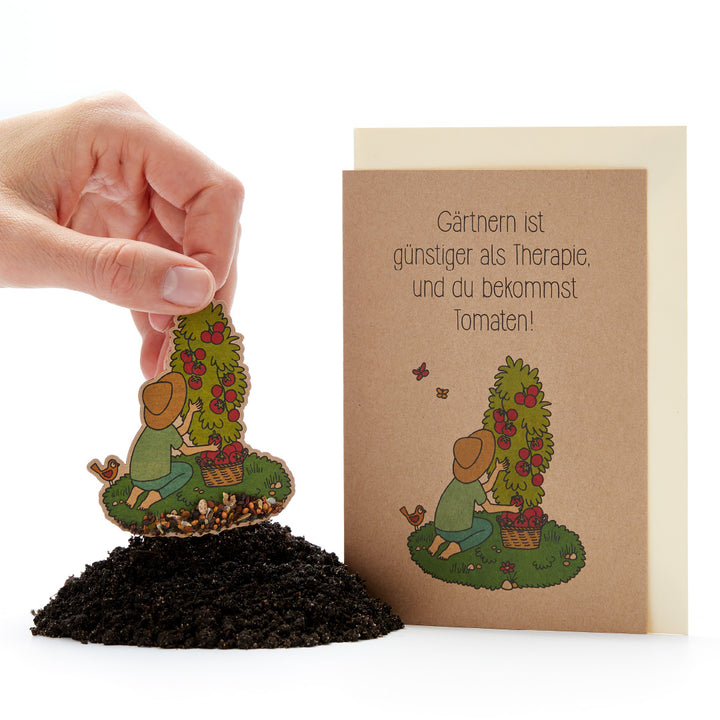 Une main plante une carte de graines représentant un jardinier et un plant de tomates dans le sol, à côté d'elle une carte de vœux sur laquelle on peut lire : « Le jardinage est moins cher que la thérapie, et vous obtenez des tomates ! »