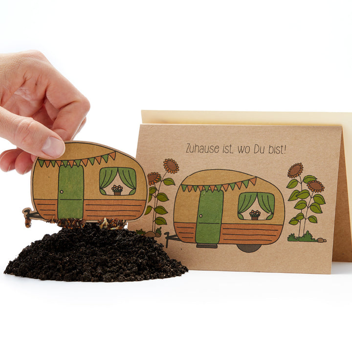 Main plaçant une carte de plantation avec une caravane illustrée dans le sol, texte « La maison est là où vous êtes ! » sur la carte"